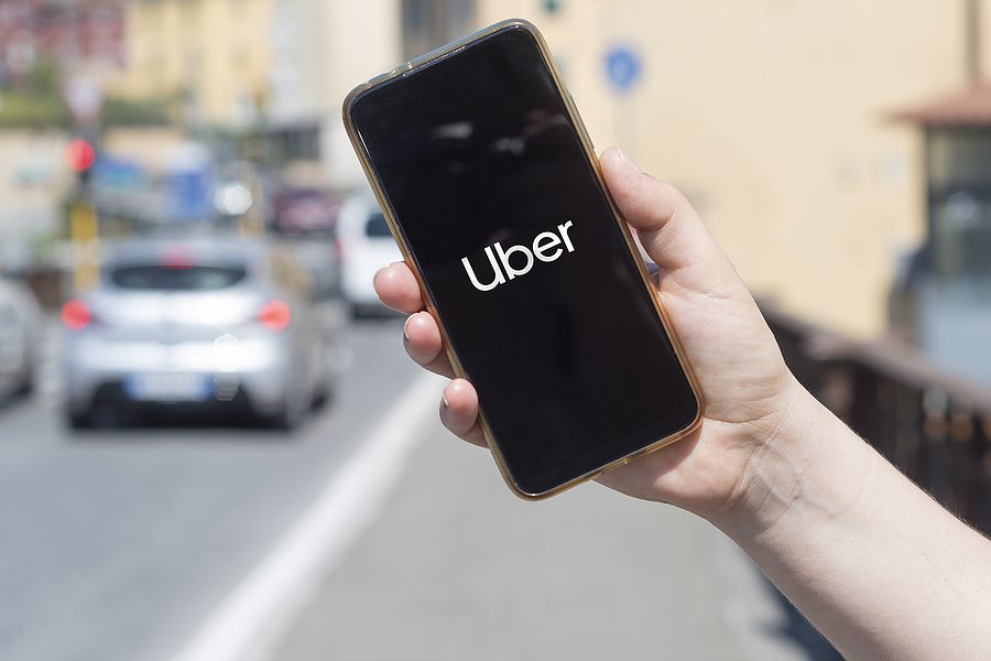 Person's hand holding a smartphone with the Uber logo on the screen with cars on a city street in the background.