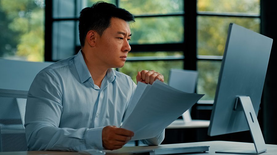 Business man sitting at desk looking concerned at computer while holding papers
