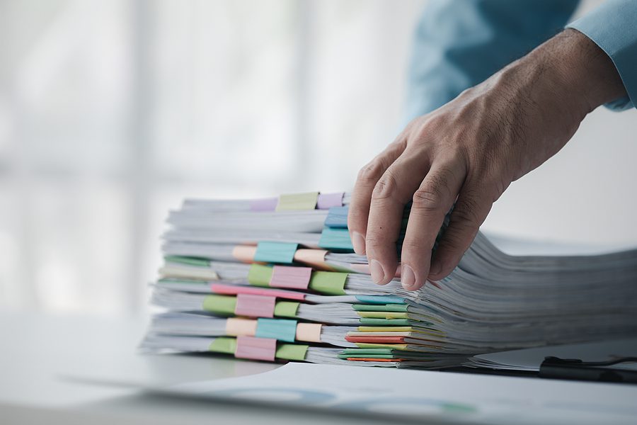 A person in an office with a huge stack of papers, he is a company employee, managing paperwork and finance, corporate filing. Document management concept.