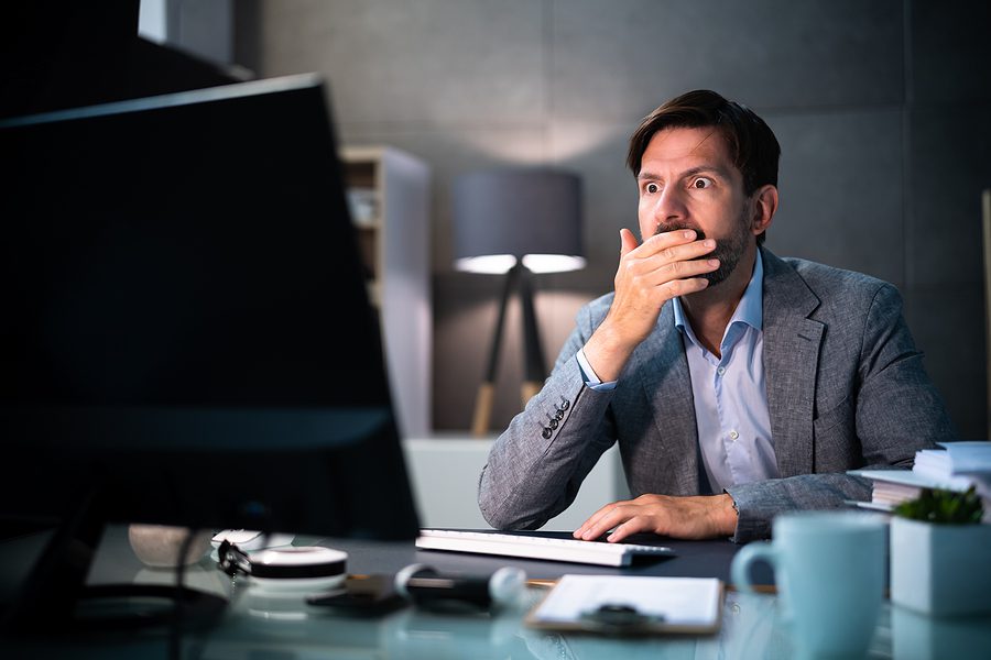 Male businessman in suit looking at his computer screen in shock with his hand over his mouth. 