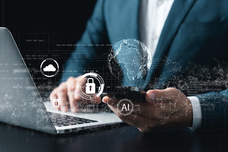 Close up of a man in a suit, holding his phone in one hand and typing on a laptop with cybersecurity concepts on the screen. 