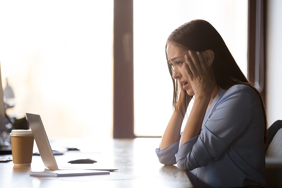 Female sitting at her desk in her office, holding her hands up to her face with a shocked look. 