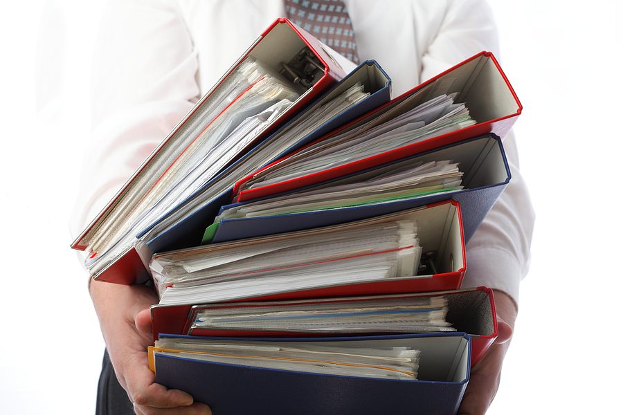 Closeup of a businessman holding a large stack of binders full of paper documents. 