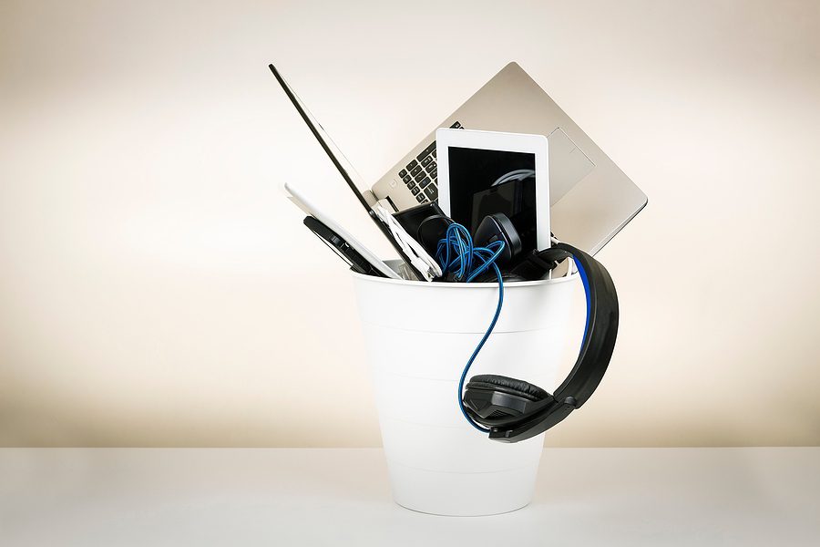 E-waste concept with small white trash can against a white backdrop filled with old electronics, including laptop, headphones, and tablets. 